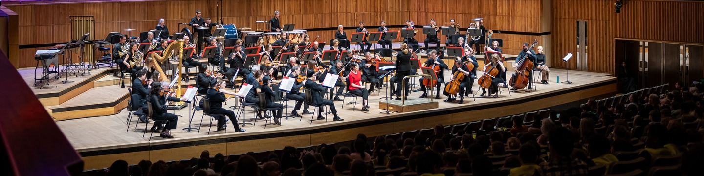 An orchestra playing on-stage at the Southbank Centre Festival Hall, watched by an audience of young people from schools in Southwark